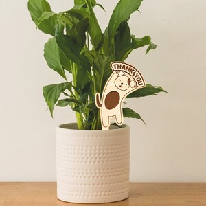 May include: A white ceramic planter with a textured surface holds a green peace lily plant. A wooden cutout of a dog holding a sign that says "Thank You" is placed in the plant.