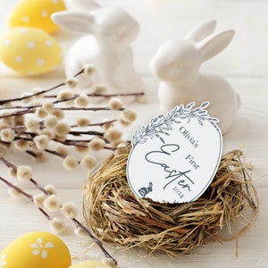 May include: A white egg-shaped card with the text "Olivia's First Easter 2024" in a floral wreath design. The card is sitting in a brown woven nest with two white ceramic bunnies and yellow Easter eggs in the background.