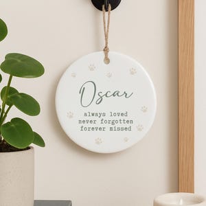 May include: A round, white ceramic memorial ornament with the name "Oscar" in elegant script. The ornament features paw prints and the text "always loved, never forgotten, forever missed." It hangs from a jute twine.