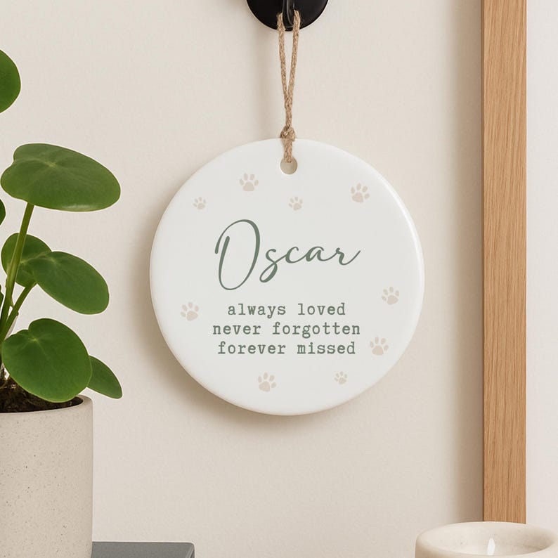 May include: A round, white ceramic memorial ornament with the name "Oscar" in elegant script. The ornament features paw prints and the text "always loved, never forgotten, forever missed." It hangs from a jute twine.