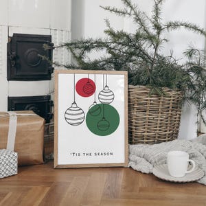 K&ouml;nnte beinhalten: Ein gerahmter Druck mit wei&szlig;em Hintergrund und einer Schwarzwei&szlig;illustration von Weihnachtskugeln, die an F&auml;den h&auml;ngen. Die Kugeln sind rot, gr&uuml;n und wei&szlig;. Der Text "'Tis the season" ist unter den Kugeln gedruckt.