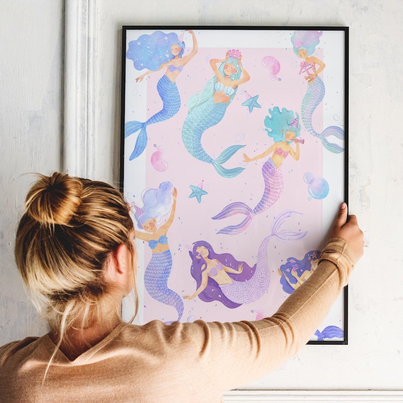 May include: A framed art print featuring watercolor illustrations of mermaids in various poses. The mermaids have colorful hair and tails, set against a light pink background with stars and bubbles. The frame is black.