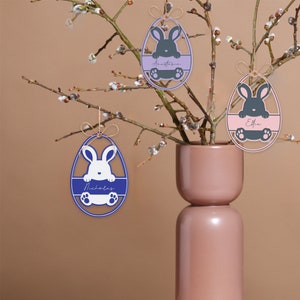 May include: Easter egg ornaments in blue, purple, and pink, each featuring a bunny design and a name. The ornaments are hanging from branches in a pink vase. The names on the ornaments are Nicholas, Anastasia, and Effie.