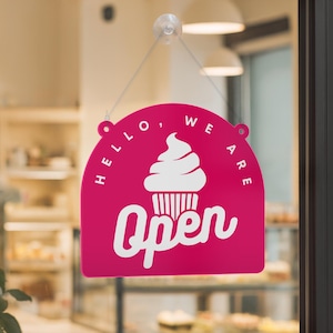 Może przedstawiać: Magenta znak w kształcie łuku z białymi napisami "Hello, We Are" i "Open". Biała grafika babeczki znajduje się na środku. Znak wisi na szklanym oknie.
