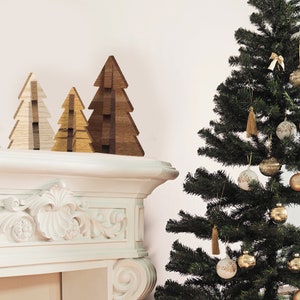 May include: Three wooden Christmas trees of varying sizes, with a brown, beige, and light brown finish, are arranged on a white mantelpiece. A larger green Christmas tree with gold ornaments and tassels is in the background.