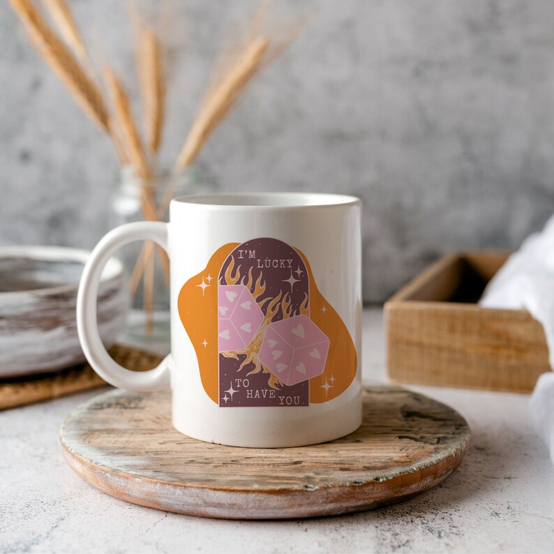 May include: White ceramic mug with a graphic of two dice with hearts on them, surrounded by flames and stars. The text "I'm Lucky to Have You" is printed on the mug.
