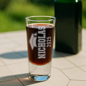 May include: A clear shot glass filled with a dark liquid, featuring the name "NICHOLAS" and the year "2025" in white lettering, along with a graduation cap graphic. A dark green bottle is blurred in the background.