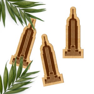 May include: Three brown wooden cutout decorations shaped like the Empire State Building. The decorations are on a white background with a green palm leaf.