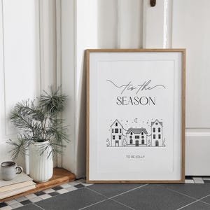 May include: A black and white printable art print with the text "Tis the season to be jolly" and a simple illustration of three houses with a crescent moon and stars in the sky.