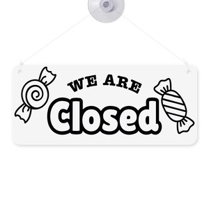 Puede incluir: Cartel rectangular blanco con las palabras "WE ARE Closed" en negro, con ilustraciones de caramelos a cada lado. El cartel est&aacute; sujeto a una ventosa transparente con una cuerda blanca.