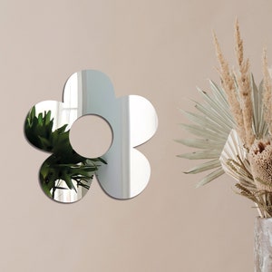 May include: A silver mirrored flower-shaped wall decor with a round center hole. The mirror reflects a green plant and a window.
