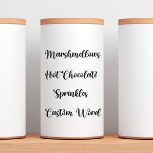 Peut inclure: Trois contenants cylindriques blancs avec des couvercles en bois. Le contenant du milieu porte une inscription en noir qui dit "Marshmallows", "Chocolat chaud", "Sprinkles" et "Mot personnalis&eacute;".