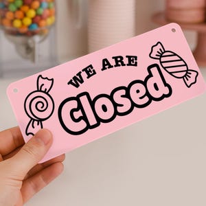 Puede incluir: Un letrero rectangular rosa con las palabras "WE ARE Closed" en negro. El letrero tiene dos ilustraciones de caramelos y dos peque&ntilde;os agujeros. El letrero est&aacute; sostenido por una mano.