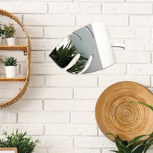 Pu&ograve; includere: Uno specchio argentato a forma di foglia di monstera, appeso a un muro di mattoni bianchi. Lo specchio riflette una pianta in vaso e uno scaffale in vimini.