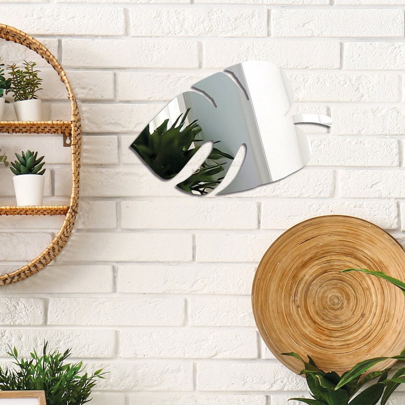Pu&ograve; includere: Uno specchio argentato a forma di foglia di monstera, appeso a un muro di mattoni bianchi. Lo specchio riflette una pianta in vaso e uno scaffale in vimini.