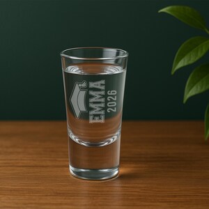 May include: A clear shot glass filled with liquid, featuring the name "EMMA" and the year "2026" etched on the front, along with a graduation cap design. The glass is set on a wooden surface.