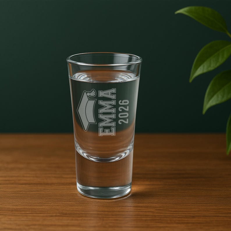 May include: A clear shot glass filled with liquid, featuring the name "EMMA" and the year "2026" etched on the front, along with a graduation cap design. The glass is set on a wooden surface.