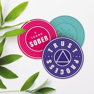 May include: Three round, colourful coasters with inspirational messages. The pink coaster says "5 Years Sober", the blue coaster says "6 Months Sober", and the purple coaster says "Trust The Process".