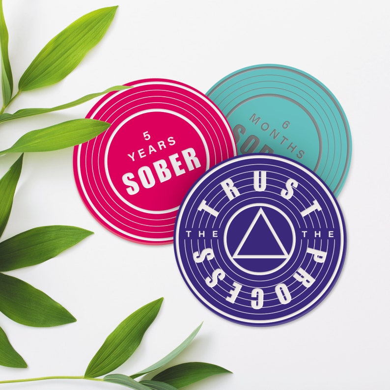 May include: Three round, colourful coasters with inspirational messages. The pink coaster says "5 Years Sober", the blue coaster says "6 Months Sober", and the purple coaster says "Trust The Process".