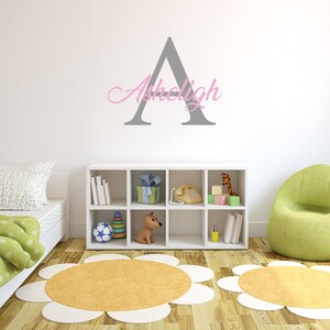 May include: A gray letter A with the name "Ashleigh" written in pink cursive above it. The letter and name are on a white wall. There is a white bookshelf with toys and books on it. There are two yellow and white flower-shaped rugs on the floor.