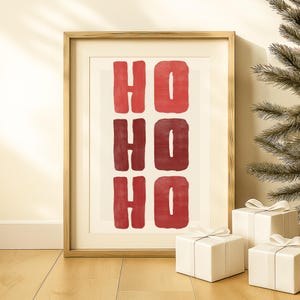May include: Framed art print with the words "HO HO HO" in red block letters. The artwork is in a gold frame and is displayed with three wrapped white gift boxes with white ribbons. A Christmas tree is in the background.