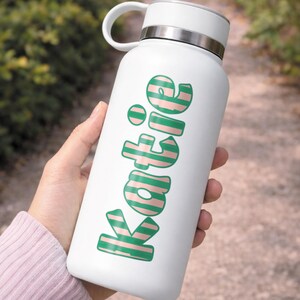 Op de afbeelding: Witte waterfles met een zilveren rand en handvat. De naam "Katie" is gedrukt in groene letters met roze strepen. De fles wordt vastgehouden door een persoon voor een vage buitenachtergrond.