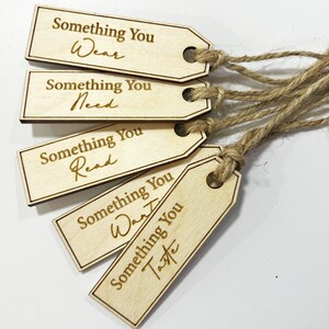 Pu&ograve; includere: Cinque etichette regalo in legno con spago attaccato. Ogni etichetta ha una frase diversa scritta su di essa: "Something You Wear", "Something You Need", "Something You Read", "Something You Want" e "Something You Taste".