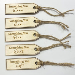 Pu&ograve; includere: Cinque etichette regalo in legno con spago attaccato. Ogni etichetta ha il testo "Something You" seguito da una parola diversa: "Wear", "Need", "Read", "Want" e "Taste".
