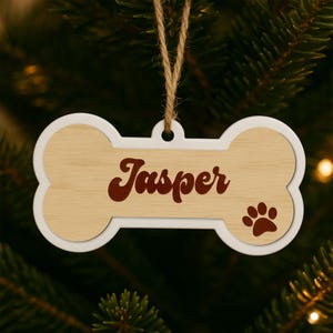 Pode incluir: Um enfeite de Natal em forma de osso com um centro de gr&atilde;o de madeira clara e uma borda branca. O nome "Jasper" est&aacute; escrito em um script vermelho escuro, e uma pegada est&aacute; no lado direito. Um la&ccedil;o de barbante est&aacute; no topo para pendurar.
