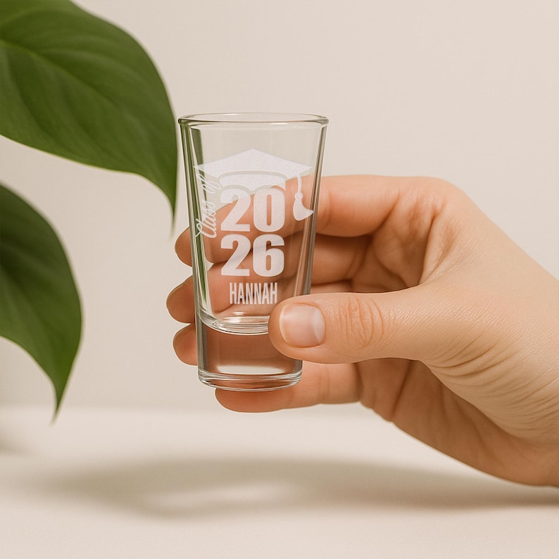 May include: Clear shot glass with white text and a graduation cap design. The text includes the numbers "2026" and the name "Hannah". The glass is held by a hand.