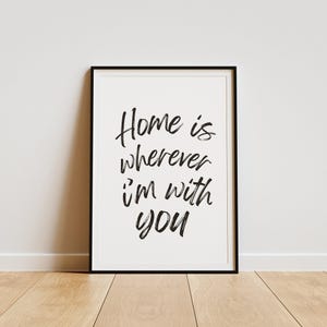 May include: Black and white typography print with the text "Home is wherever I'm with you" in a modern calligraphy style. The print is framed in a black frame and is displayed on a light wood floor.