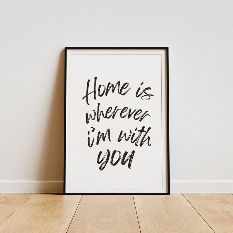 May include: Black and white typography print with the text "Home is wherever I'm with you" in a modern calligraphy style. The print is framed in a black frame and is displayed on a light wood floor.
