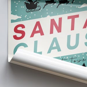 K&ouml;nnte beinhalten: Ein Weihnachtsposter im Vintage-Stil mit dem Text "SANTA CLAUS" in roten und t&uuml;rkisfarbenen Buchstaben auf hellblauem Hintergrund mit wei&szlig;en Schneeflocken. Das Poster hat einen wei&szlig;en Rand und eine teilweise abgezogene Ecke.