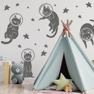 Pode incluir: Uma tenda tipi azul claro com um padr&atilde;o de estrelas cinza e branco no interior. O tipi est&aacute; instalado em um quarto com uma parede cinza e adesivos de astronautas gatos cinza. Os adesivos s&atilde;o de gatos que usam capacetes de astronautas e flutuam no espa&ccedil;o.