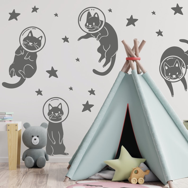 Pode incluir: Uma tenda tipi azul claro com um padr&atilde;o de estrelas cinza e branco no interior. O tipi est&aacute; instalado em um quarto com uma parede cinza e adesivos de astronautas gatos cinza. Os adesivos s&atilde;o de gatos que usam capacetes de astronautas e flutuam no espa&ccedil;o.