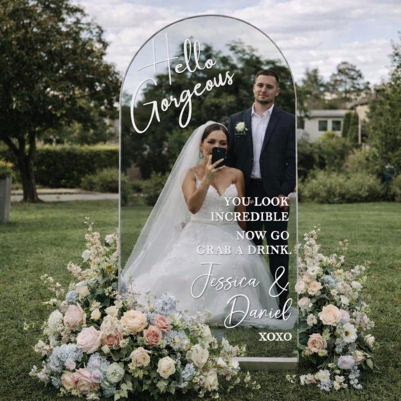 May include: A clear acrylic wedding sign with an arched top, featuring the words "Hello Gorgeous" and a couple. The sign includes the text "You look incredible, now go grab a drink," and the names "Jessica & Daniel" with "xoxo" at the bottom. Floral arrangements frame the sign.