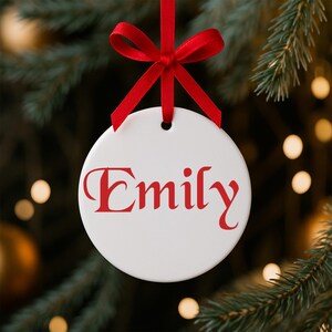 May include: A white, round Christmas ornament with the name "Emily" in red script. The ornament hangs from a red satin ribbon bow against a blurred background of a Christmas tree.
