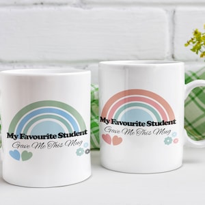 May include: Two white ceramic mugs with rainbow designs and the text "My Favourite Student Gave Me This Mug". The mugs are on a white surface with a green and white checkered cloth.