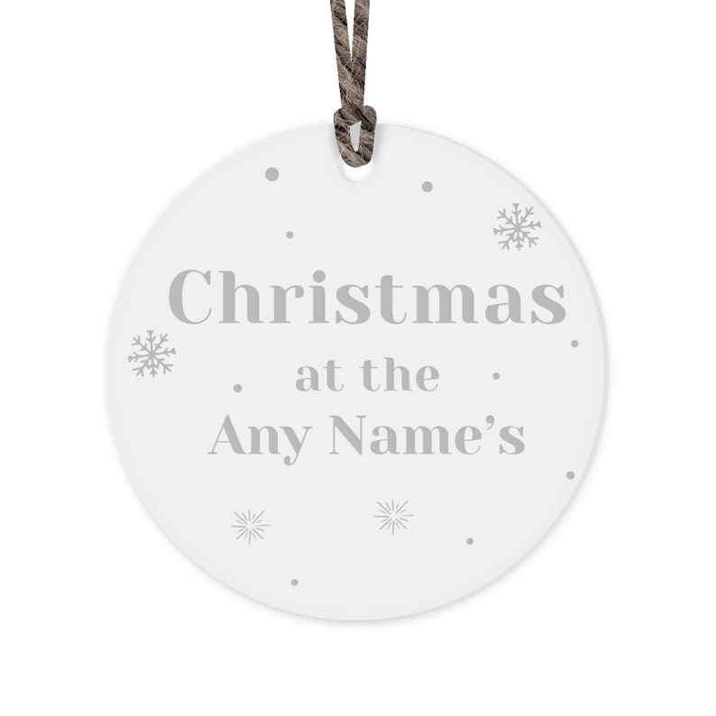 Peut inclure: Ornement de No&euml;l rond blanc avec l'inscription "Christmas at the Any Name's" en gris. L'ornement pr&eacute;sente des motifs de flocons de neige et de points, et une ficelle marron pour l'accrocher.