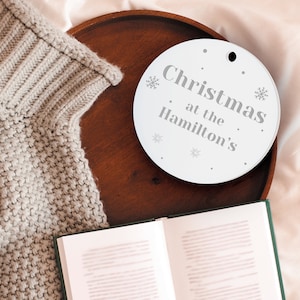 Peut inclure: Ornement de No&euml;l rond blanc avec l'inscription "Christmas at the Hamilton's" en gris. L'ornement est orn&eacute; de flocons de neige et d'un trou pour l'accrocher. Il repose sur un plateau en bois fonc&eacute;, &agrave; c&ocirc;t&eacute; d'un pull en tricot gris et d'un livre ouvert.