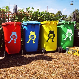 May include: Four colorful trash bins: red, blue, yellow, and green. Each bin has a frog graphic with the number "201". The bins are on wheels and are in front of a backdrop of green foliage and a blue sky.
