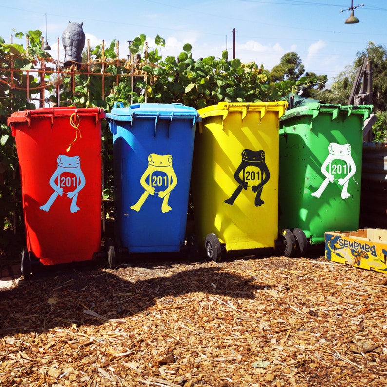 May include: Four colorful trash bins: red, blue, yellow, and green. Each bin has a frog graphic with the number "201". The bins are on wheels and are in front of a backdrop of green foliage and a blue sky.