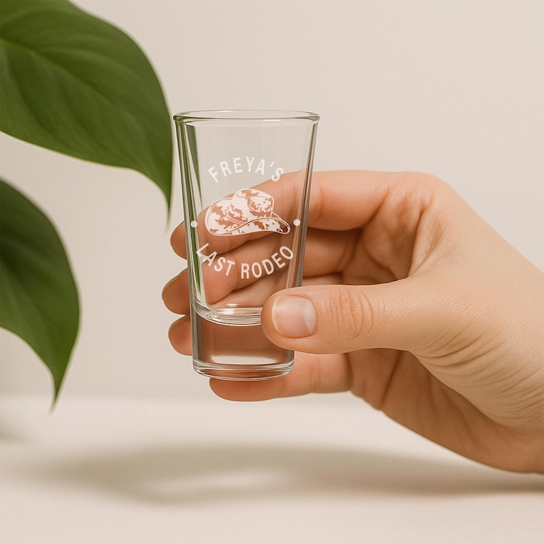 K&ouml;nnte beinhalten: Ein klares Schnapsglas mit dem wei&szlig;en Text "FREYA'S LAST RODEO". Das Design beinhaltet eine Cowboyhut-Illustration. Das Glas wird von einer Hand gehalten, mit einem weichen Hintergrund.