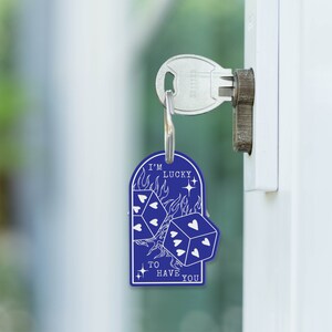 Puede incluir: Llavero azul con un dise&ntilde;o blanco de dos dados con corazones en los lados y llamas detr&aacute;s de ellos. El texto "I'm Lucky to Have You" est&aacute; escrito debajo de los dados.