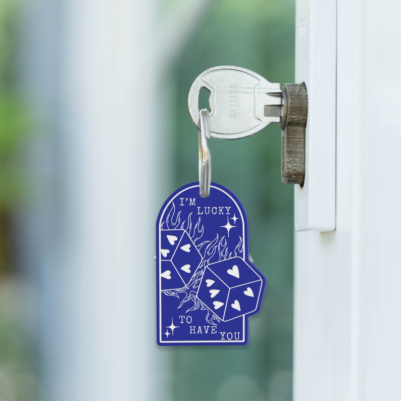Puede incluir: Llavero azul con un dise&ntilde;o blanco de dos dados con corazones en los lados y llamas detr&aacute;s de ellos. El texto "I'm Lucky to Have You" est&aacute; escrito debajo de los dados.