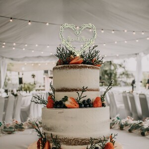 May include: A three-tiered wedding cake decorated with fresh strawberries and blueberries. The cake is topped with a wooden heart-shaped topper that says "Mr. & Mrs."