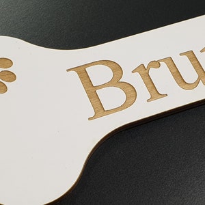 May include: White wooden bone-shaped sign with the name "Bruno" engraved in brown lettering. A brown paw print with a heart in the center is also engraved on the sign.