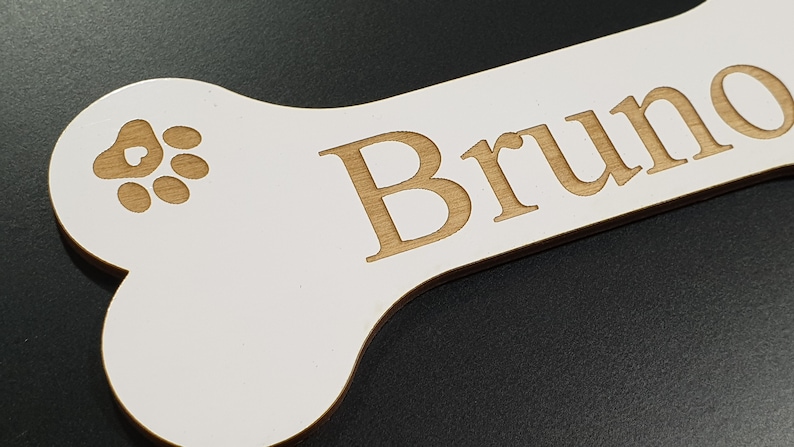 May include: White wooden bone-shaped sign with the name "Bruno" engraved in brown lettering. A brown paw print with a heart in the center is also engraved on the sign.