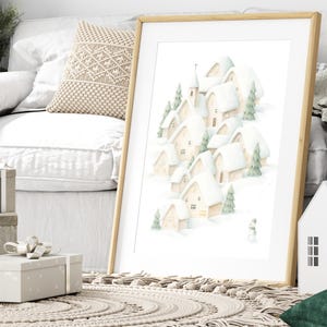 May include: A framed print showing a watercolour illustration of a snow-covered village. The artwork depicts several houses, a church, and evergreen trees, all blanketed in white snow. A small snowman is also visible. The frame is made of light-coloured wood.