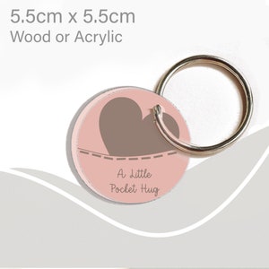 May include: A pink and brown round keychain with a heart design and the text "A Little Pocket Hug".